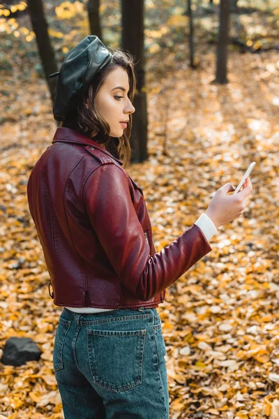 Vista lateral de la mujer joven en boina y chaqueta de cuero elegante usando teléfono inteligente en el bosque - foto de stock