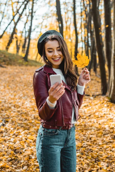 Sorridente ragazza elegante in giacca di pelle prendendo selfie con foglia gialla su smartphone all'aperto — Foto stock