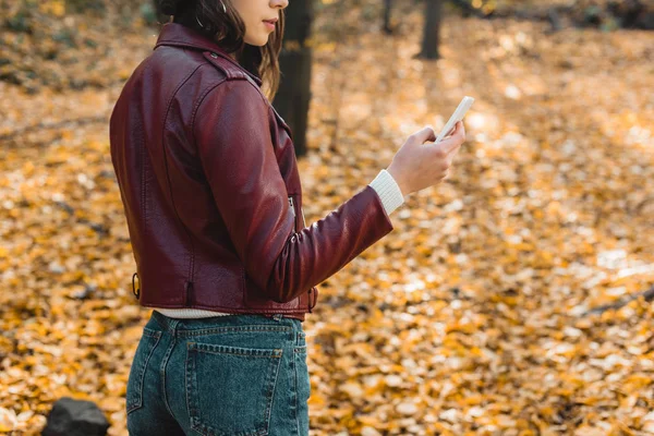 Vista parcial de mujer joven en chaqueta de cuero con estilo usando teléfono inteligente en bosque otoñal - foto de stock