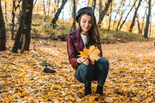 Glückliches stilvolles Mädchen in Baskenmütze und Lederjacke sitzt mit Gelb im herbstlichen Wald — Stockfoto