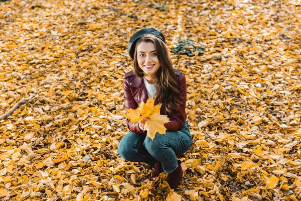 Vue grand angle de fille heureuse à la mode en béret et veste en cuir assis avec jaune dans la forêt automnale — Stock Photo