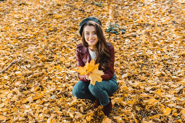 High angle view of happy fashionable girl in beret and leather jacket sitting with yellow in autumnal forest — Stock Photo