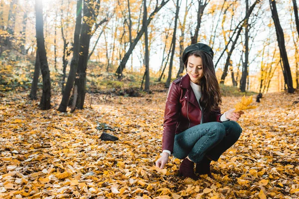 Schöne junge Frau in stylischer Baskenmütze und Lederjacke, die im herbstlichen Wald gelbe Blätter sammelt — Stockfoto