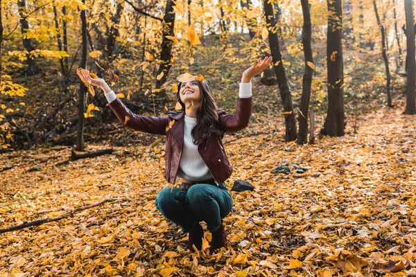 Smiling attractive woman in stylish leather jacket having fun with yellow leaves in autumnal forest — Stock Photo