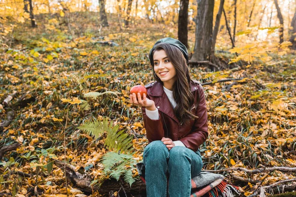Fille élégante souriante en veste en cuir assis sur la couverture et tenant pomme rouge dans la forêt — Photo de stock