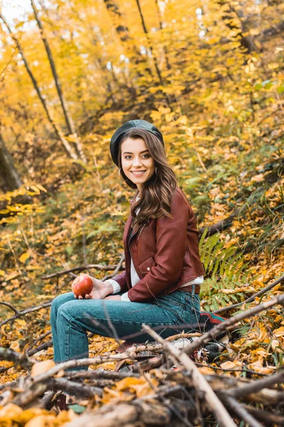 Femme élégante souriante en béret et veste en cuir tenant pomme et regardant la caméra à l'extérieur — Photo de stock