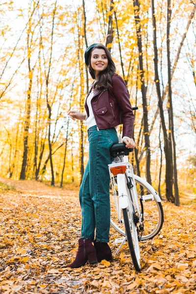 Low angle view of happy female model in beret and leather jacket posing near bicycle in autumnal forest — Stock Photo