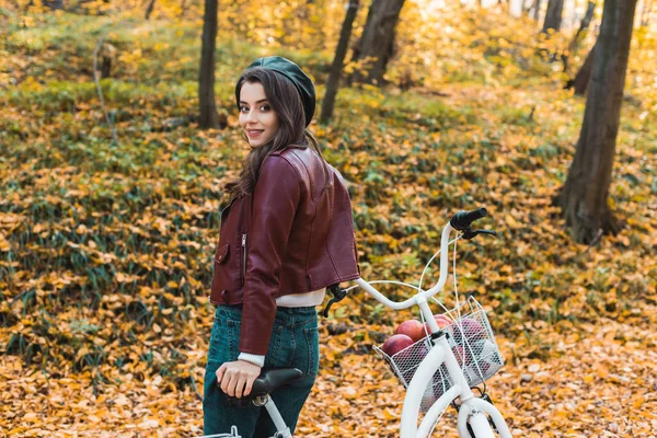 Selective focus of fashionable female model in beret and leather jacket posing near bicycle in autumnal forest — Stock Photo
