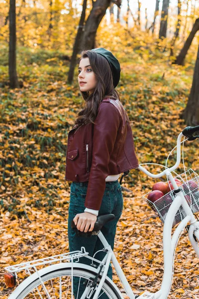 Jolie fille élégante en béret et veste en cuir posant près de vélo dans la forêt automnale — Photo de stock