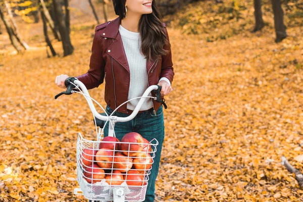 Vue partielle de la jeune fille à la mode en veste en cuir portant vélo avec panier plein de pommes rouges dans le parc automnal — Stock Photo