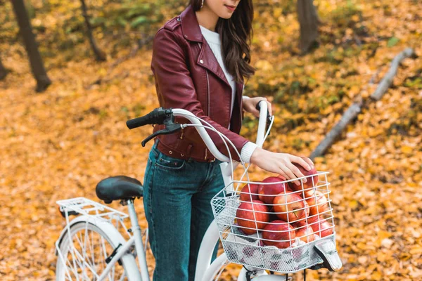 Cropped image of stylish woman in leather jacket taking apple from basket of bicycle outdoors — Stock Photo