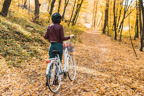 Visão traseira da mulher em jaqueta de couro elegante e boina transportando bicicleta na floresta outonal — Fotografia de Stock