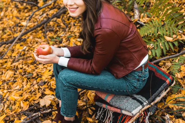 Partial view of stylish girl in leather jacket sitting on blanket and holding red apple in autumnal forest — Stock Photo