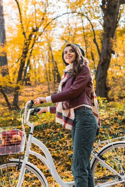 Low angle view of smiling woman in stylish leather jacket and beret carrying bicycle in autumnal forest — Stock Photo