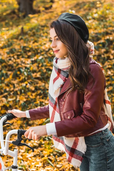 Foco seletivo da mulher alegre na jaqueta de couro elegante e boina transportando bicicleta ao ar livre — Fotografia de Stock