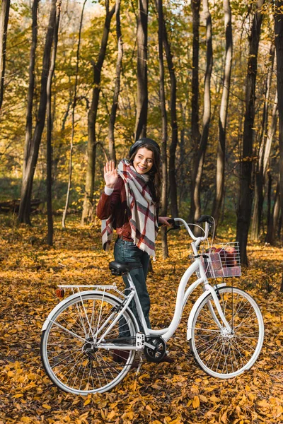 Улыбающаяся стильная девушка в кожаной куртке и берете машет рукой и несет велосипед в осеннем парке — стоковое фото