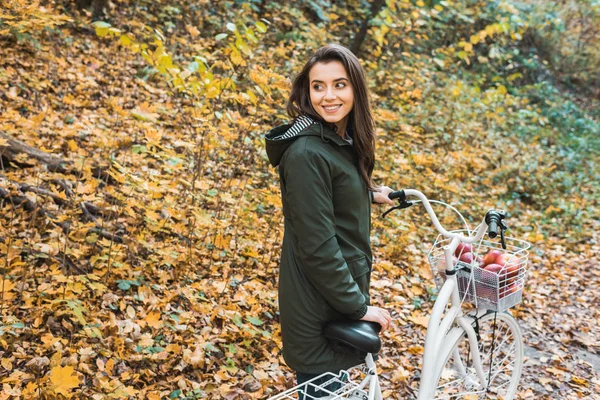 Lächelnde junge Frau trägt Fahrrad mit Korb voller Äpfel im gelben Herbstwald — Stockfoto