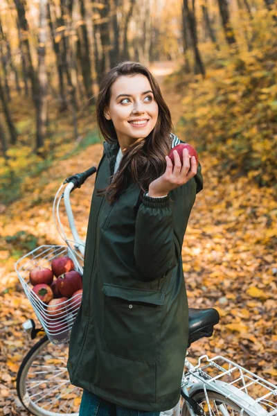 Attrayant jeune femme avec pomme debout près de vélo dans la forêt automnale — Photo de stock