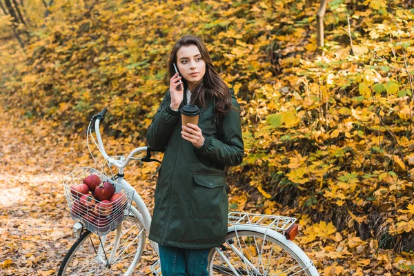 Mujer joven con taza de café de papel hablando en el teléfono inteligente cerca de la bicicleta en el bosque otoñal - foto de stock