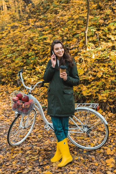Jeune femme souriante avec tasse de café en papier parlant sur smartphone près de vélo dans la forêt automnale — Photo de stock