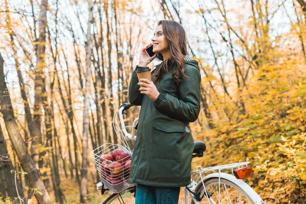 Vue à angle bas de femme heureuse avec tasse de café parlant sur smartphone près de vélo dans la forêt automnale — Photo de stock