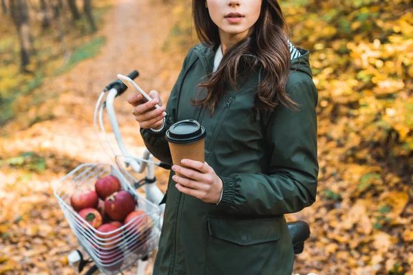 Обрізане зображення жінки з одноразовою чашкою кави, використовуючи смартфон біля велосипеда на відкритому повітрі — стокове фото