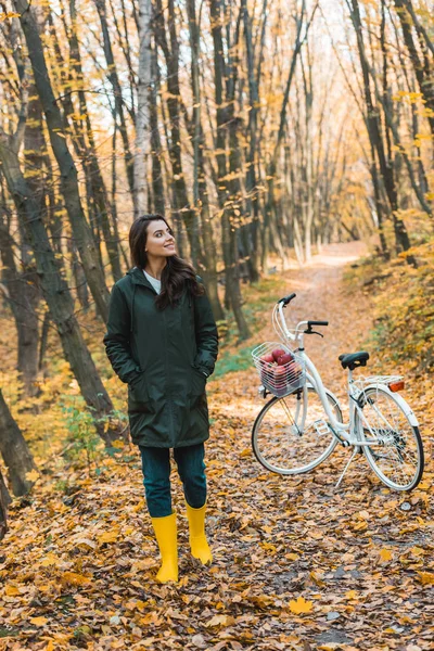 Chica feliz en botas de goma amarillas caminando cerca de la bicicleta en el bosque otoñal - foto de stock