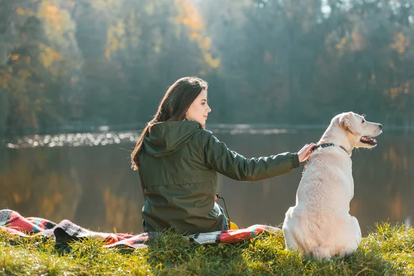 Rear view of young woman siting on blanket with adorable golden retriever near pond in park — Stock Photo
