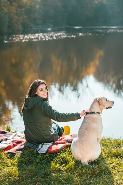 Smiling young woman siting on blanket with adorable golden retriever near pond in park — Stock Photo