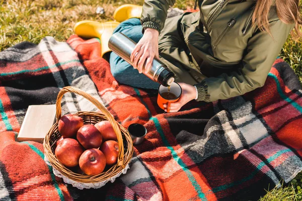 Partial view of woman pouring coffee from thermos into cup while sitting on blanket with basket full of apples outdoors — Stock Photo