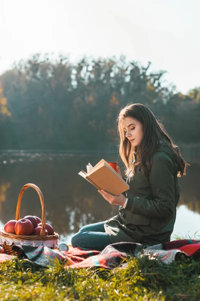 Vista laterale di attraente giovane donna con libro di lettura tazza di caffè sulla coperta vicino stagno nel parco — Foto stock