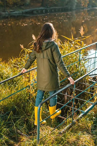 Rear view of woman in jacket and yellow rubber boots posing near pond in park — Stock Photo