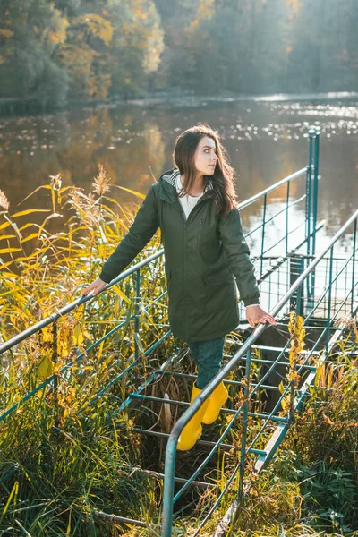 Вид сбоку молодой женщины в куртке и желтых резиновых сапогах, позирующей возле пруда в парке — стоковое фото