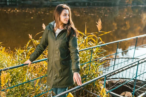 Jeune femme souriante en veste posant près de l'étang dans le parc — Photo de stock