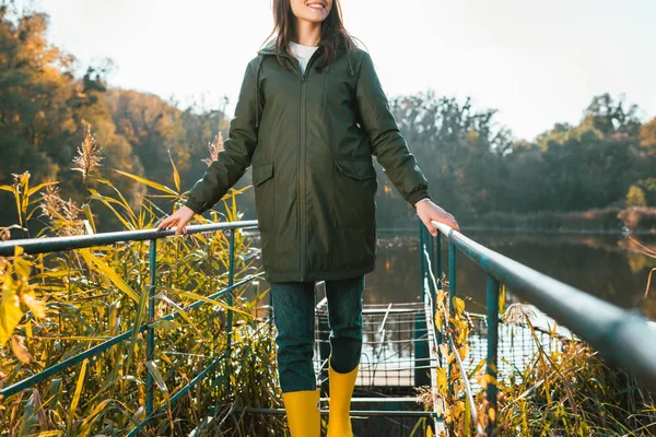 Partial view of young woman in jacket and yellow rubber boots posing near pond in park — Stock Photo