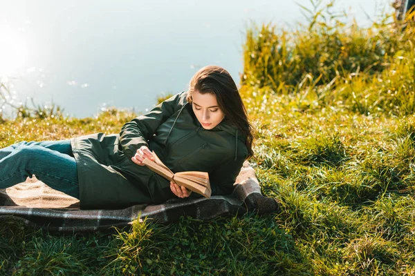 Belle femme lecture livre tout en posant sur la couverture près de l'étang dans le parc — Photo de stock