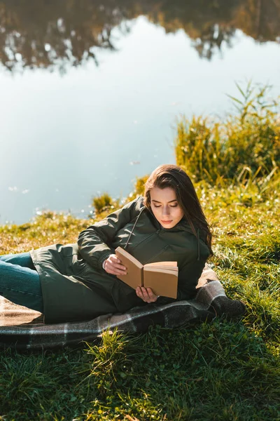 Vista de ángulo alto de la mujer joven leyendo libro mientras que pone en manta cerca del estanque en el parque - foto de stock