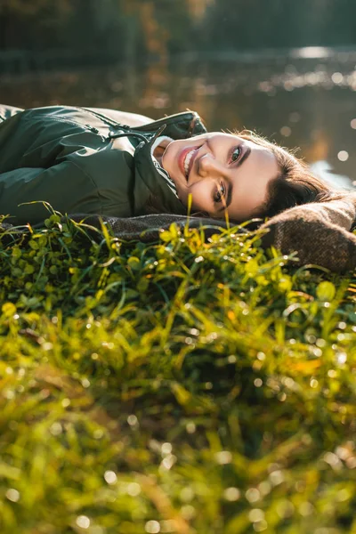 Selektiver Fokus einer lächelnden Frau, die sich auf eine Decke legt und im Freien in die Kamera blickt — Stockfoto