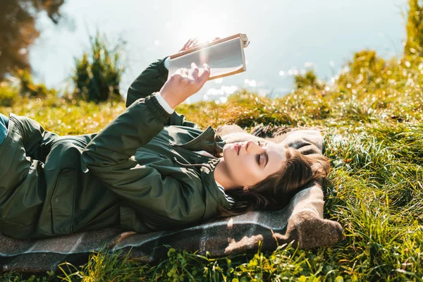 Side view of cheerful young woman reading book while laying on blanket near pond in park — Stock Photo