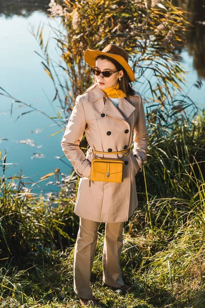 High angle view of fashionable woman in hat and trench coat posing near pond in park — Stock Photo