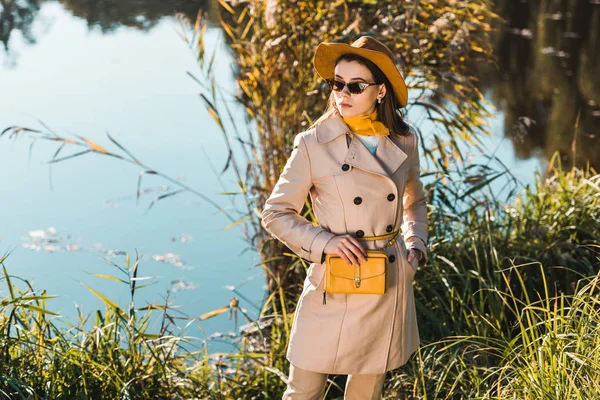 Elegante Frau in Trenchcoat und Hut posiert mit gelber Tasche im Freien — Stockfoto
