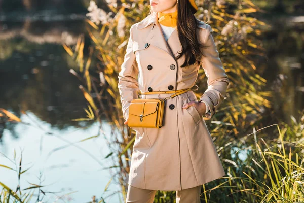 Частичный вид стильной женщины в плаще, позирующей с желтой сумкой возле пруда в парке — стоковое фото