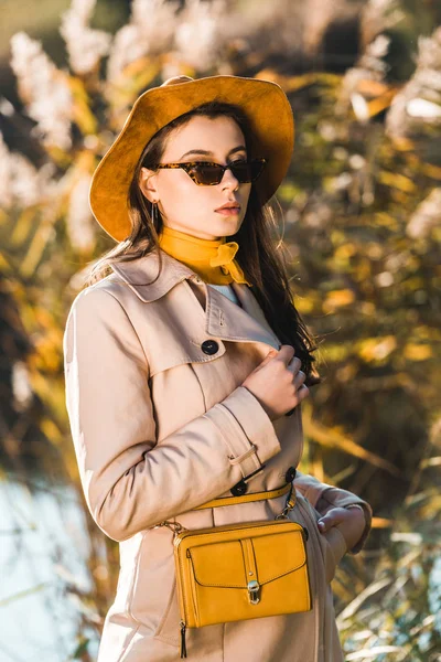 Attrayant femme en trench coat posant avec sac jaune à l'extérieur — Photo de stock