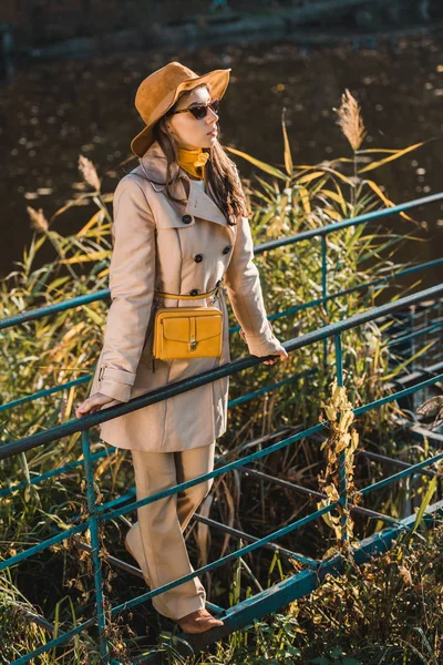 Belle jeune femme en lunettes de soleil, trench coat et chapeau posant près de rambarde — Photo de stock