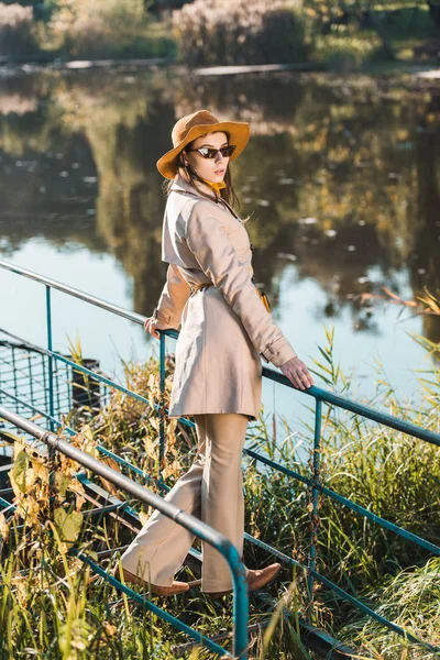 Selective focus of stylish female model in sunglasses, trench coat and hat posing near pond in park — Stock Photo