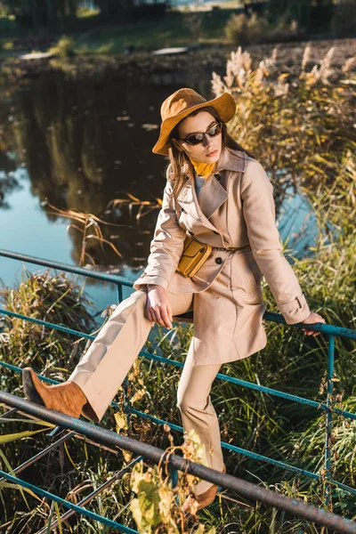 Attractive woman in sunglasses, trench coat and hat sitting on railing near pond in park — Stock Photo