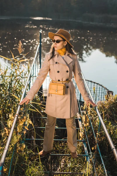 Giovane donna alla moda in occhiali da sole, trench e cappello in posa vicino stagno nel parco — Foto stock