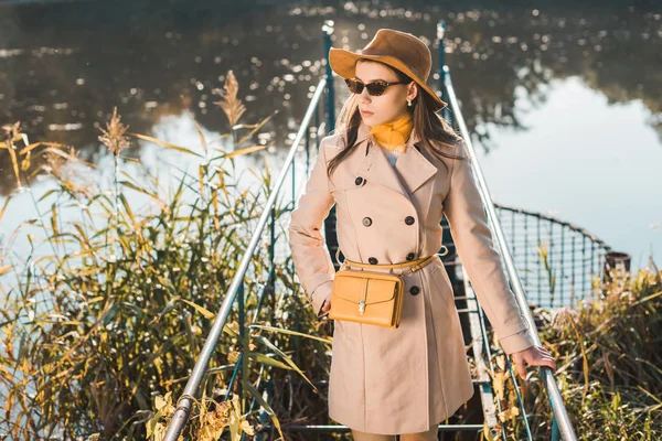 Modische Frau mit Sonnenbrille, Trenchcoat und Hut posiert am Teich im Park — Stockfoto