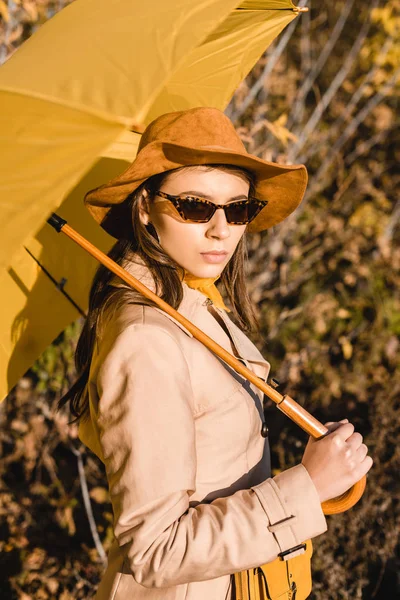 Élégante femme élégante en lunettes de soleil et chapeau regardant la caméra et tenant parapluie jaune à l'extérieur — Photo de stock