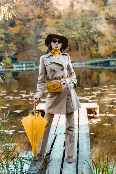 Mujer con estilo de confianza en gafas de sol, gabardina y sombrero posando con paraguas amarillo cerca del estanque en el parque - foto de stock